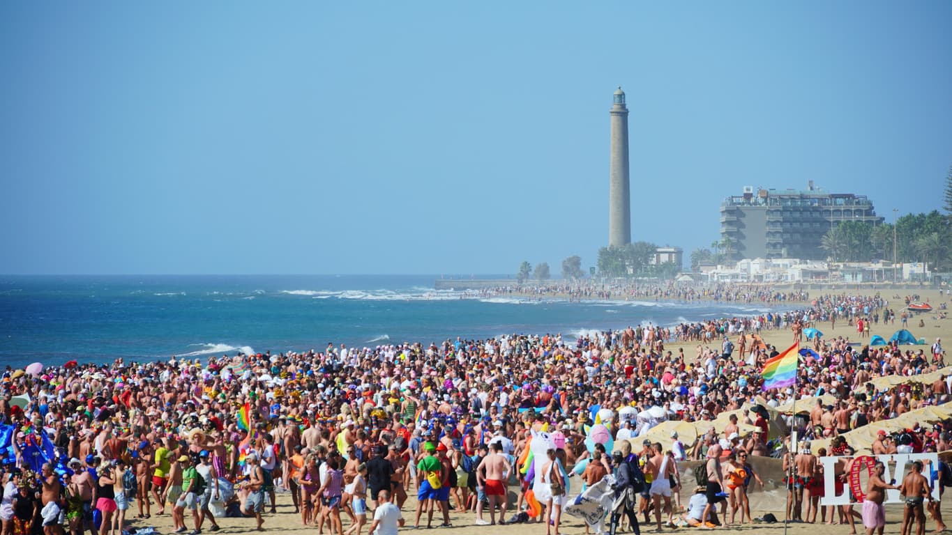 A crowd in LBGTQ+ colors on Maspalomas beach, with the iconic lighthouse in the background.
