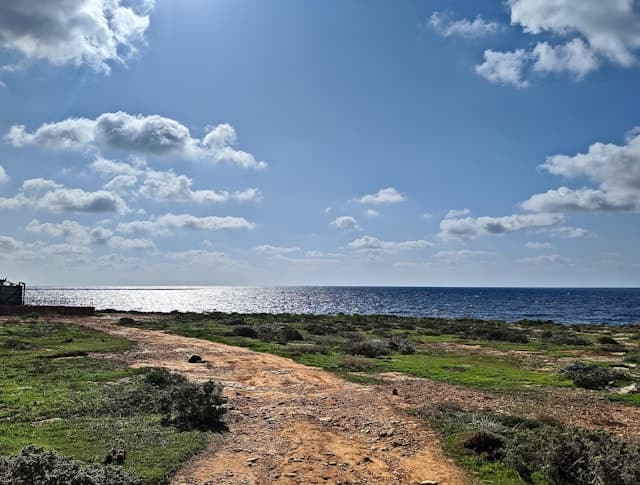 Sentiero sterrato con vista sul mare a Lampedusa, sotto un cielo azzurro con nuvole sparse.