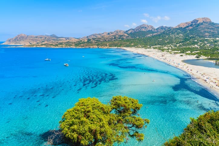 Veduta panoramica della spiaggia di Ostriconi in Corsica, con mare turchese, sabbia chiara e rilievi montuosi sullo sfondo.