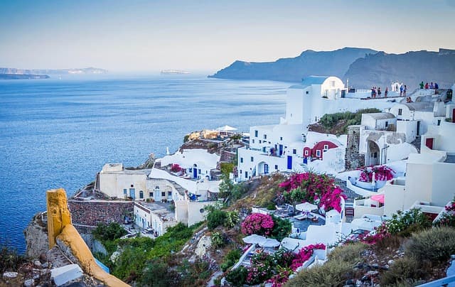 Vista panoramica di Oia a Santorini con case bianche e cupole blu, bouganville in fiore e il Mar Egeo sullo sfondo.