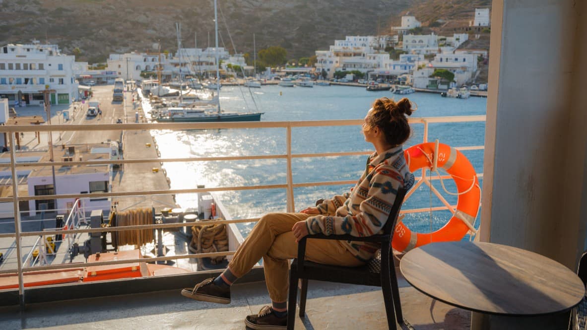 Djevojka sjedi na stolcu na palubi trajekta i gleda na obalu.