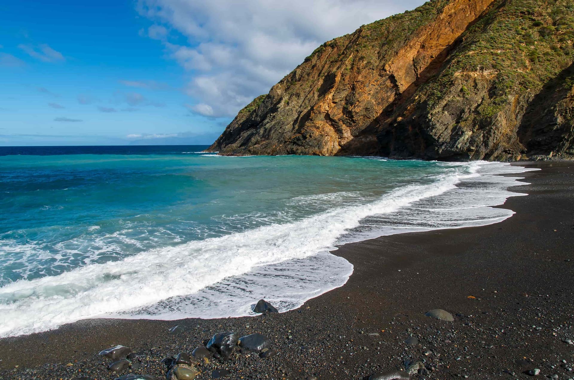 A lush cliff in front of a beach with black sand and turquoise water