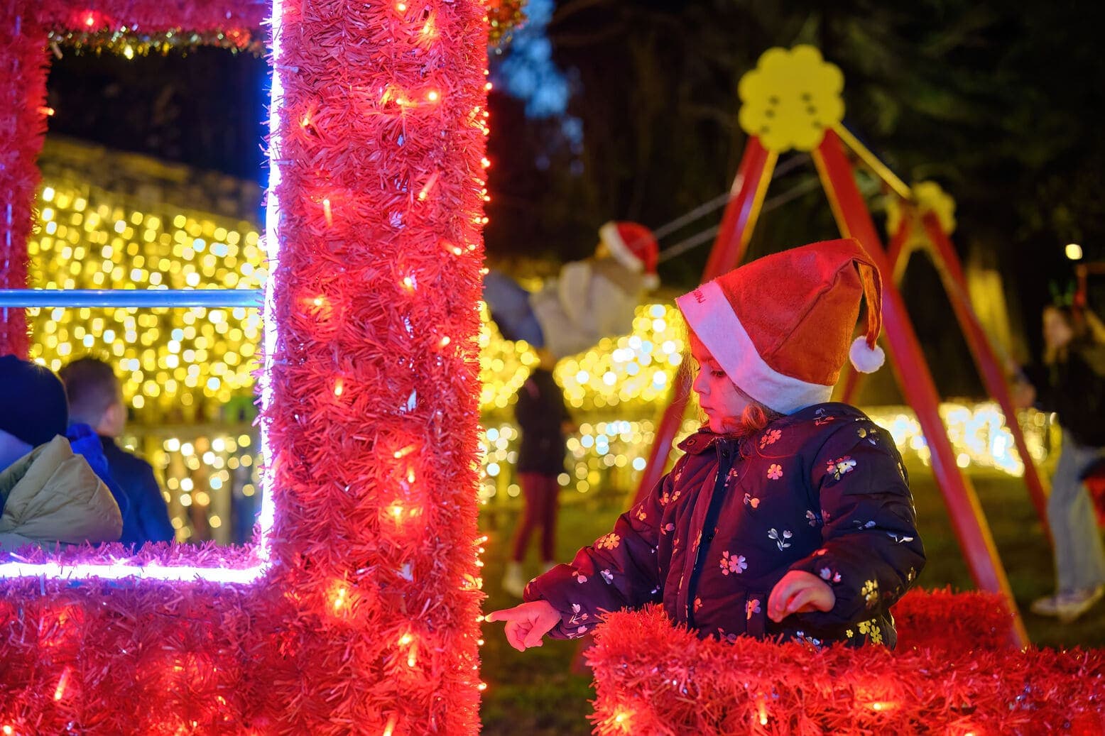 Djevojčica sa božićnom kapom na glavi divi se crvenim lampicama u parku na adventu na otoku Lošinju u večernjim satima.