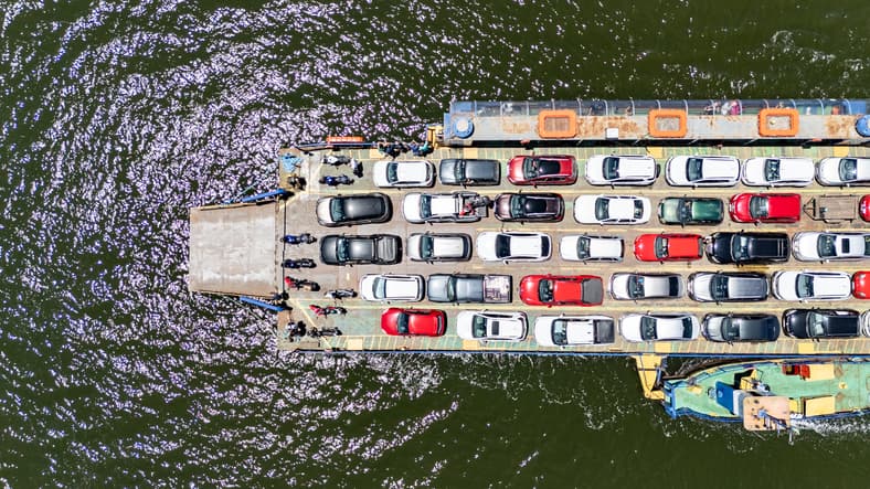 aerial view of a ferry transporting several cars, motorcycles and trucks, across a river, on a sunny day