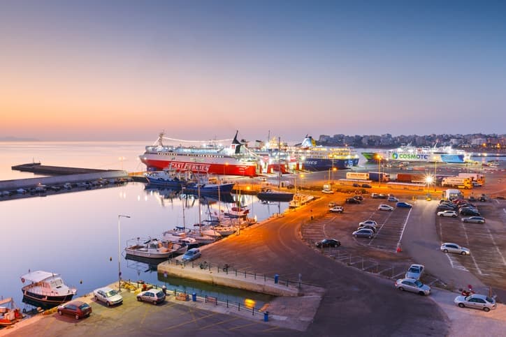 Porto greco al tramonto con traghetti ormeggiati e luci che si riflettono sull’acqua.