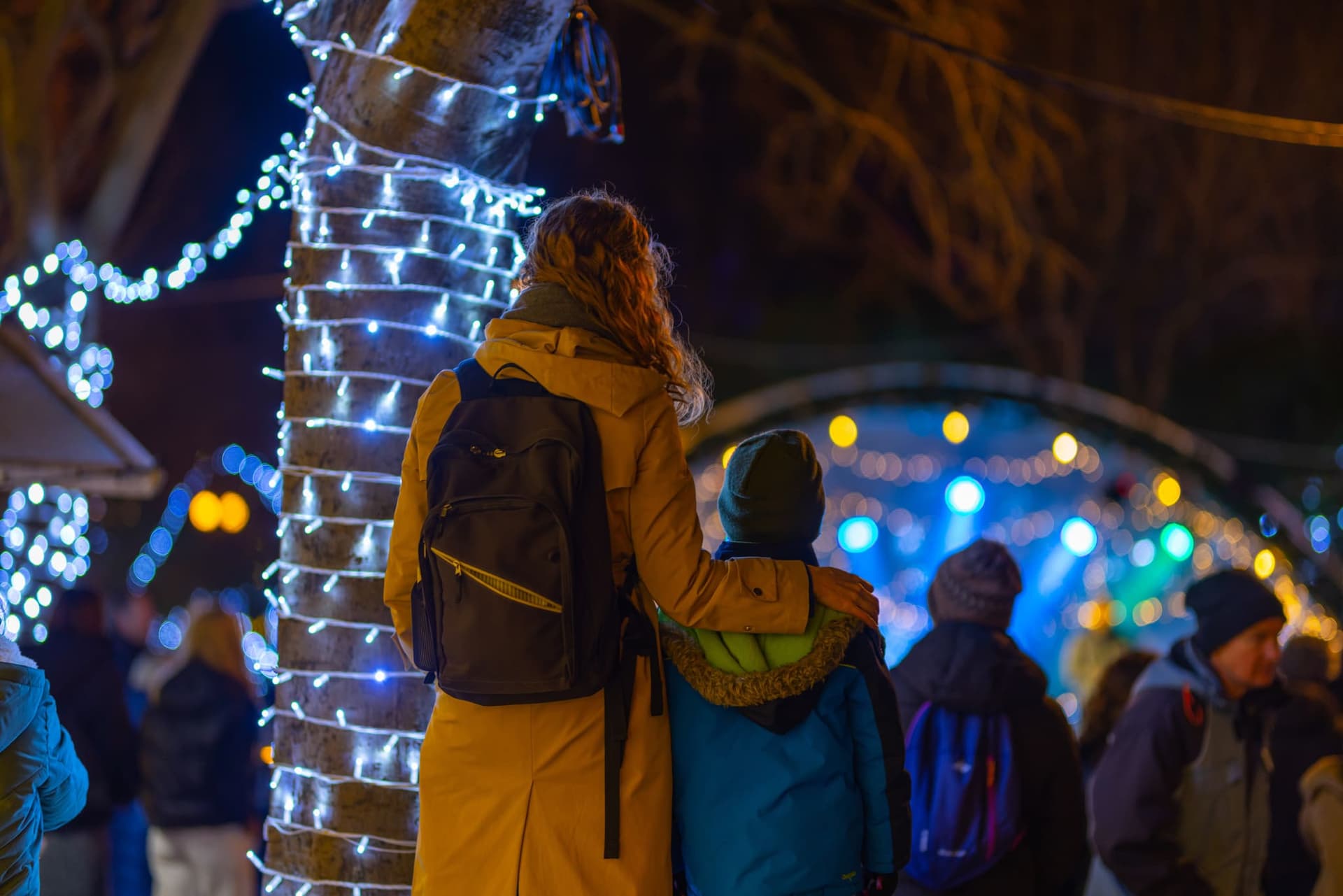 Majka grli sina desnom rukom oko ramena, oboje stoje okrenuti leđima i gledaju naprijed, ispred njih su lampice na adventu u gradu Krku na otoku Krku.