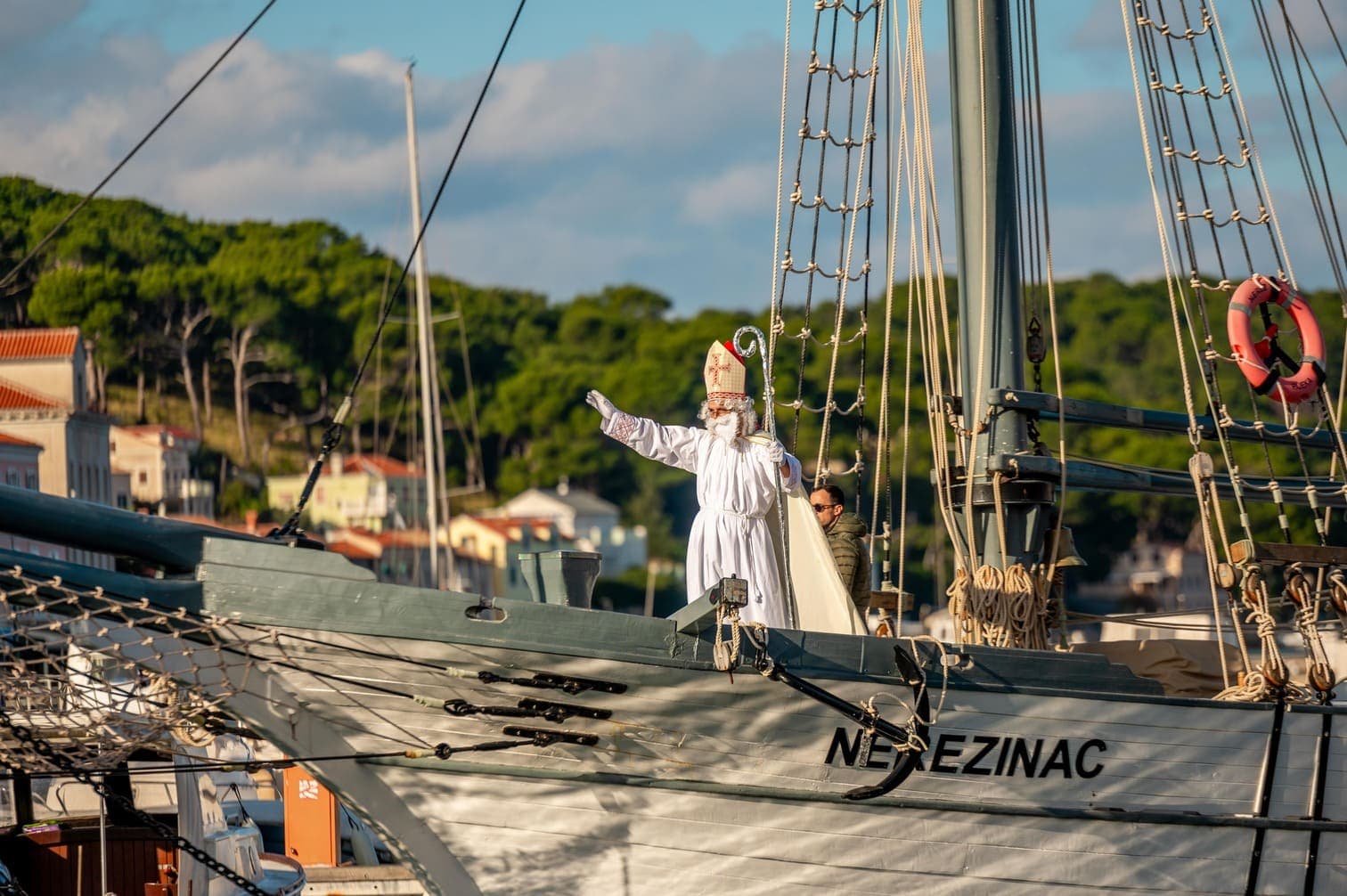Muškarac u bijelom kostimu Svetog Nikole s podignutomg rukom pozdrava uplovljava na brodu u luku na otoku Lošinju.