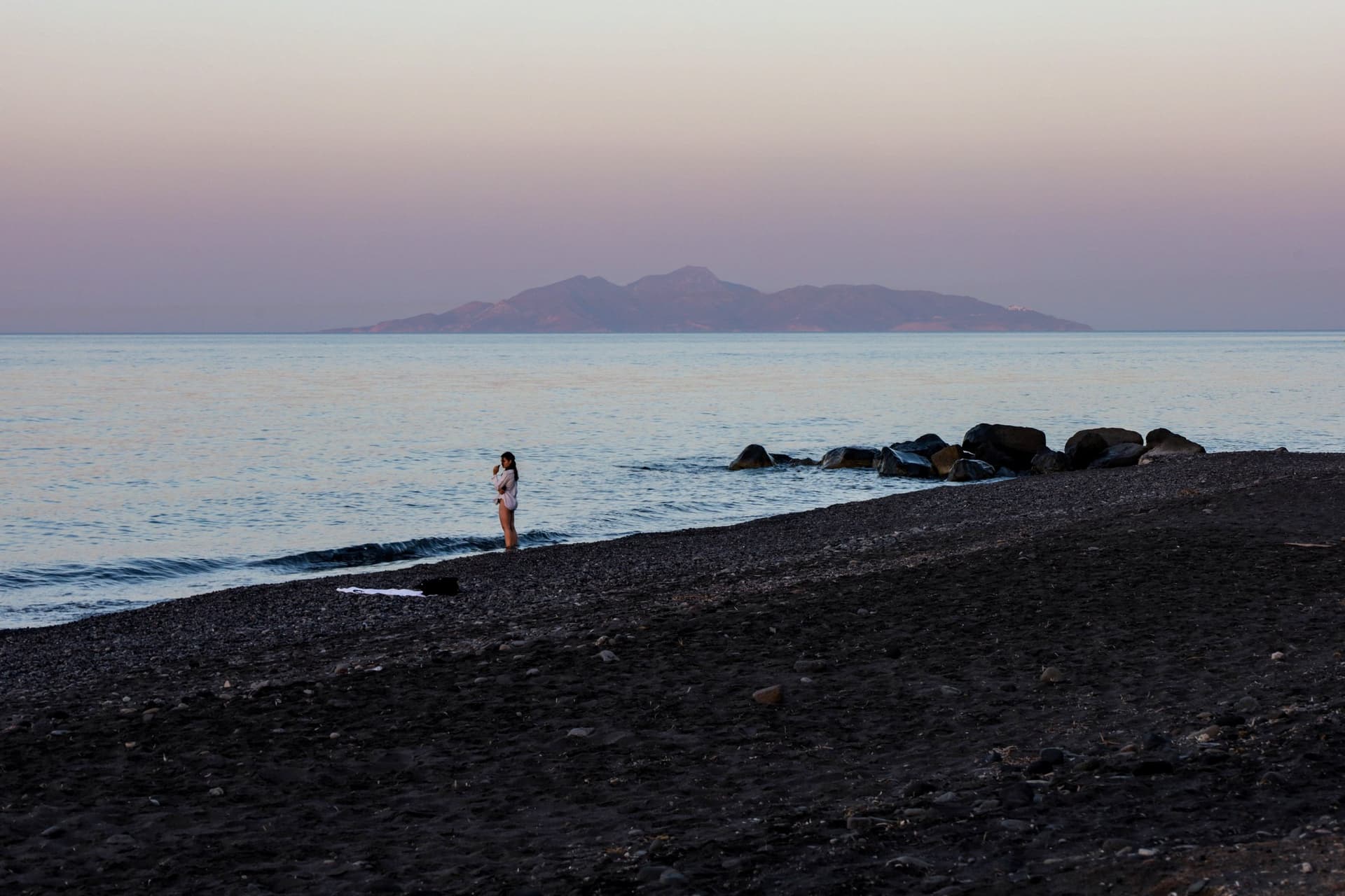A woman stands distantly on the black sand beaches of Anafi, Greece.