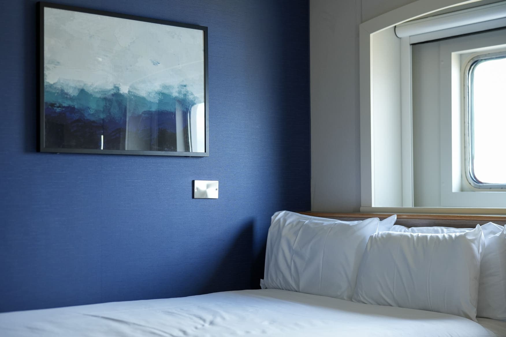 Kabina na trajektu Spirit of France kompanije P&O Ferries s bračnim krevetom, bijelom posteljinom, plavim zidom sa apstraktnom slikom i prozorom s pogledom na ocean.