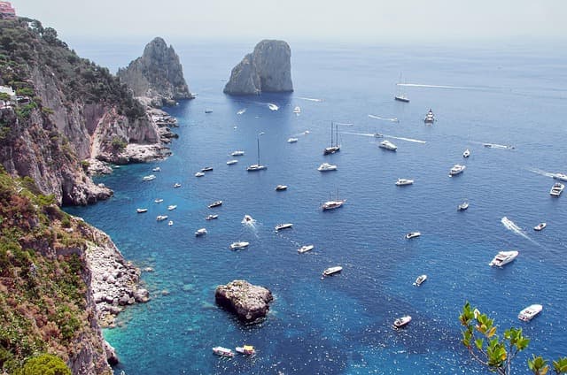 Vista dei Faraglioni di Capri con barche nel mare turchese lungo la costa rocciosa dell’isola.