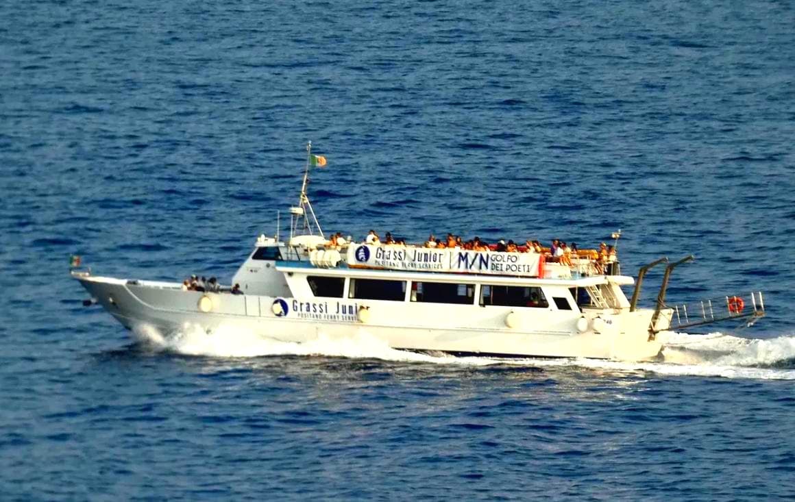 A ferry boat gliding over the blue Amalfi Coast seas with people on the outside deck.