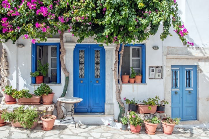 Casa tradizionale di Tinos con porta blu, facciata bianca decorata da bouganville e vasi di fiori lungo il vicolo.