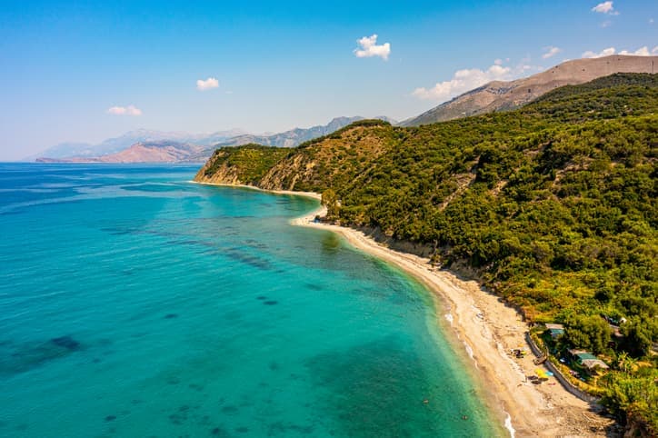 Veduta panoramica della costa albanese con spiaggia di ciottoli, mare turchese e colline verdi affacciate sul Mar Ionio.