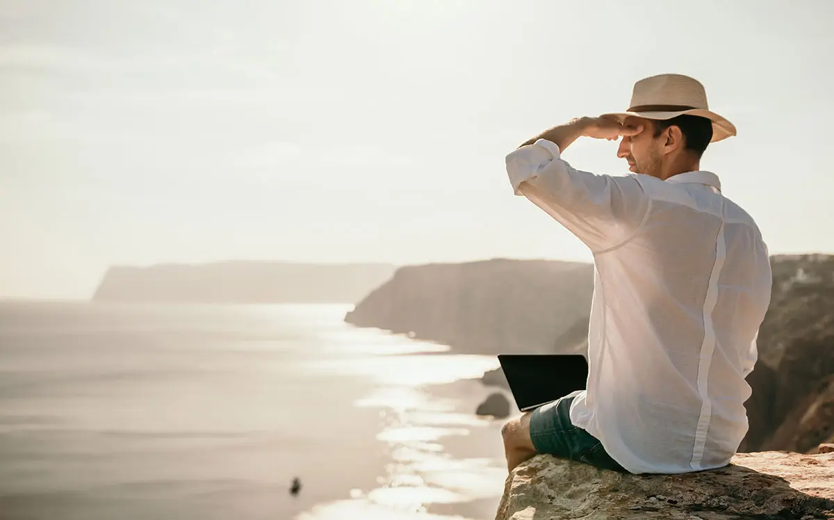 Man sitting on a cliff with a laptop, looking out over the sea near sunset.