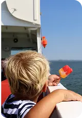 Ett blont småbarn äter en glasspinne och tittar på vågorna som passerar från färjans däck.