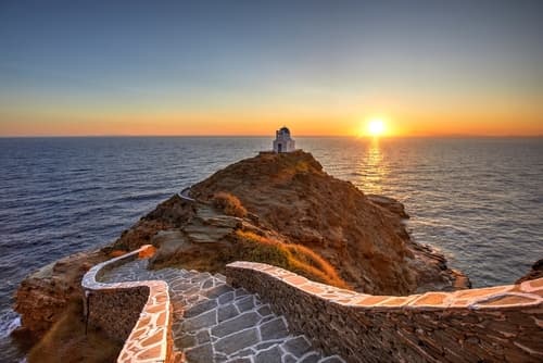 Sunset view of a winding path leading to a small white chapel on a rocky outcrop overlooking the sea.