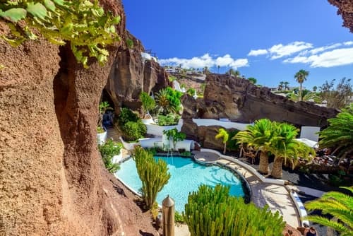 A lush oasis with a turquoise pool surrounded by tropical plants and rock formations, under a sunny sky, creating a serene and inviting natural retreat.