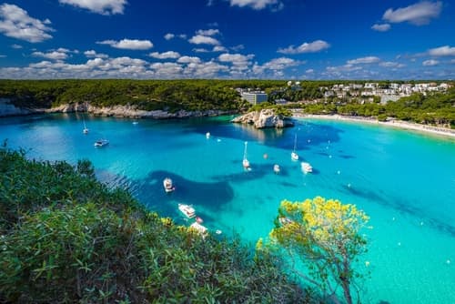 A stunning coastal scene with turquoise waters, moored boats, a secluded beach, and a backdrop of cliffs and lush vegetation under a bright blue sky with clouds.