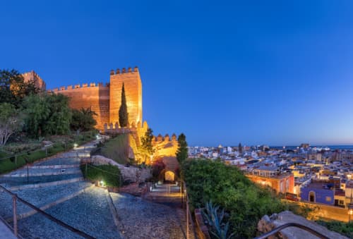 Overlooking the city of Almería, the ancient Alcazaba fortress stands on a hill, with a panoramic view of the urban landscape beneath a clear sky.