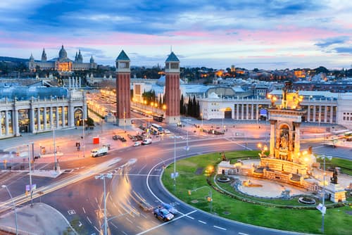 Twilight over a bustling city square with illuminated monuments, twin towers, traffic trails, and a historical building in the background under a pastel-hued sky.