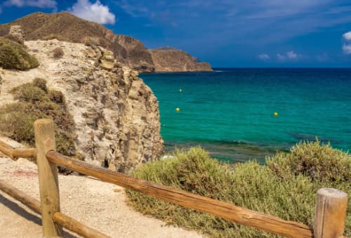 A tranquil beach in the Cabo de Gata-Níjar Natural Park, Spain, with clear turquoise waters, bordered by a rugged hillside and a wooden fence.