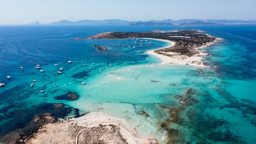 An aerial view of a breathtaking sandbar with crystal clear turquoise waters, surrounded by boats and a rugged coastline, under a clear blue sky.