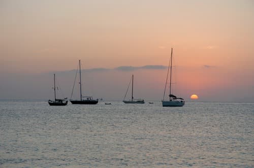 Sailboats on calm waters against a soft dusk sky, with the sun dipping below the horizon, casting a gentle orange glow over the serene seascape.