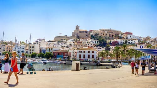 People walking along the harbor in Ibiza Town with white buildings and the historical fortress of Dalt Vila in the background under a sunny sky.