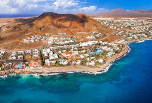 Coastal resort town by a deep blue sea, adjacent to a barren volcanic hill, with scattered white buildings under a cloudy blue sky.