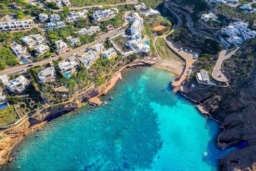 Aerial view of a turquoise cove with surrounding white buildings, winding roads, and lush greenery on a sunlit Mediterranean coastline.