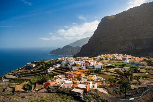 A coastal village with colorful buildings and terraced fields, nestled between a steep cliff and the sea, under a bright sky with scattered clouds.