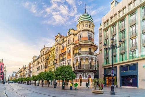 A city street at dusk with charming architecture, featuring a distinctive tower, ornate facades, and a tree-lined sidewalk leading into the distance.