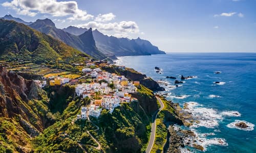 A coastal village nestled on a cliffside with lush greenery, overlooking a rugged coastline and the ocean, with dramatic mountain ridges in the background.