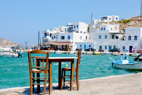Chairs facing a harbour on Tinos Island, Greece, with traditional white buildings and boats in the background, under a bright blue sky.