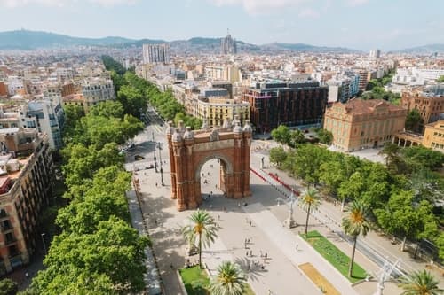 Bird's-eye view of the Arc de Triomf and promenade in Barcelona, with lush green trees, urban buildings, and a hazy skyline in the background.
