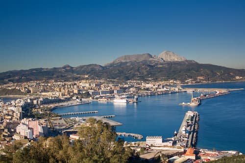 Panoramic view of Ceuta’s coastline featuring the port, Mediterranean Sea, urban landscape, and the Monte Hacho in the background under a clear blue sky.