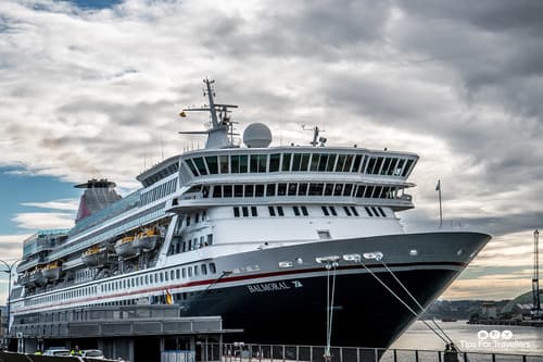 Cruise ship docked at a port, its imposing black hull and white superstructure set against a backdrop of dramatic clouds and a hint of blue sky.