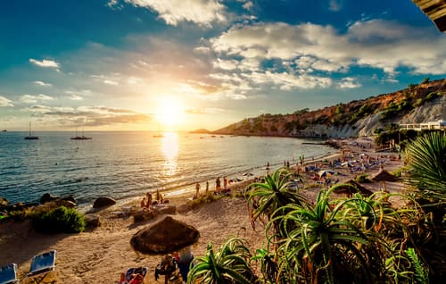Sunset at an Ibiza beach with people enjoying the evening, thatched umbrellas, and boats on the water, framed by lush greenery under a vibrant sky.