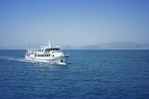A ferry cruises on the blue waters of the Mediterranean Sea towards Ibiza with a hazy mountainous coastline in the distance under a clear sky.