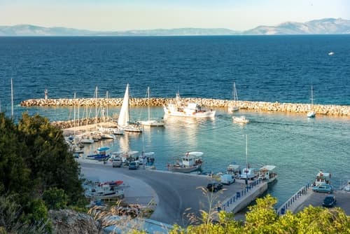 Elevated view of a serene marina in Rafina, Greece, with boats moored in the calm sea and a rugged coastline in the background during early evening.