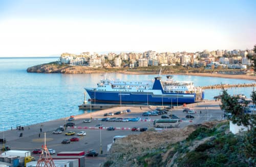 Rafina port in Greece during the golden hour, featuring a large blue ferry docked and cars lined up for boarding, with the town rising in the background.