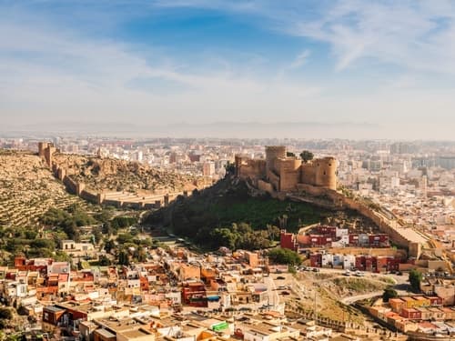 Overlooking the city of Almería, the ancient Alcazaba fortress stands on a hill, with a panoramic view of the urban landscape beneath a clear sky.