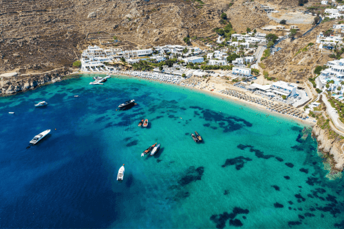 popular beach of mykonos with blue and green water while boats are on it and on the shore there are sunbeds and white houses
