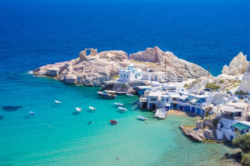 Aerial view of a small Greek cove with turquoise waters, white houses, and boats, set against a rugged landscape and open blue sea.