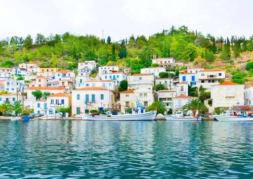 Peaceful harbor with boats and multicolored houses nestled on a verdant hillside, reflecting in the water, under an overcast sky.