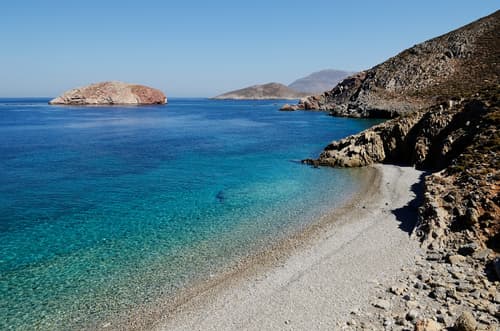 Secluded pebble beach with crystal clear azure waters, surrounded by rugged cliffs and islets under the bright Mediterranean sun.