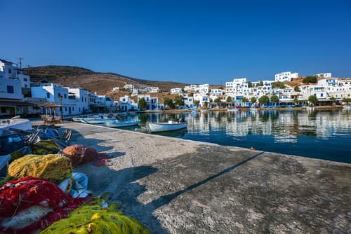 Greek fishing village with colorful nets in the foreground, white buildings with blue accents, calm waters, and a hilly backdrop under a clear sky.
