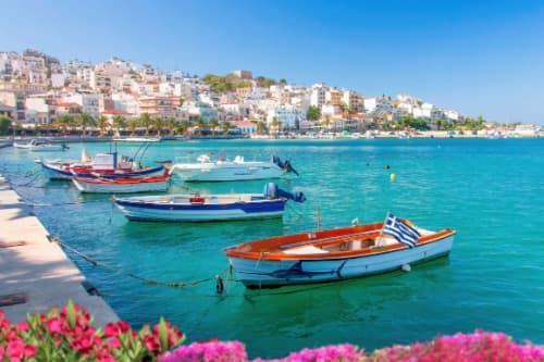 Colourful boats floating on the calm turquoise waters of Sitia's harbor, with white buildings cascading up the hillside and flowers in the foreground.