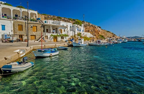 Quaint Greek harbor with clear turquoise waters, moored boats, and traditional white houses against a rugged hillside under a bright blue sky.