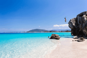 Sandstrand mit hellblauem Wasser und einem hohen Felsen am Ufer, von dem aus ein Mann ins Meer stürzt